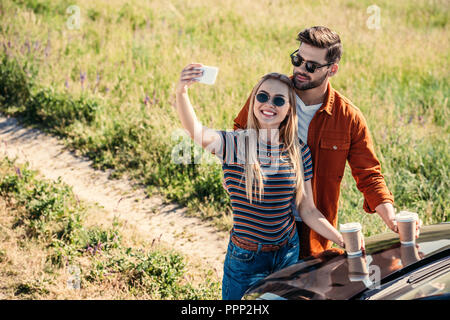 Ansicht des stilvollen Paar in Sonnenbrille mit Kaffeetassen, selfie auf dem Smartphone in der Nähe von Auto auf ländlichen Wiese Stockfoto