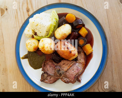 Englisch Mittagessen Abendessen essen Lammkoteletts braten Kartoffeln Blumenkohl gebratene Möhren Rotwein, Bratensoße und Minze blauen Rand weiße Platte auf einem Holztisch Stockfoto