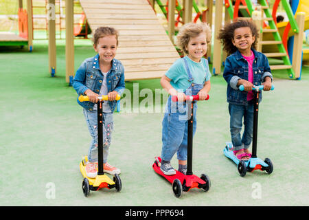 Drei multikulturellen adorable kleine Kinder reiten auf Tretroller am Spielplatz Stockfoto