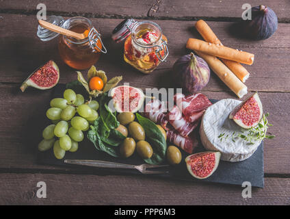 Prosciutto, Camembert, Feigen, Spinatblätter, Honig, Trauben, Oliven auf dunklen, das Brett über rustikal Hintergrund Stockfoto