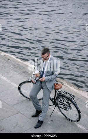 Geschäftsmann mit Kaffee mit Smartphone und stehend mit dem Fahrrad am Kai zu gehen Stockfoto