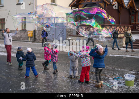 Karpacz, Polen - Februar 2018: Kinder, die versuchen, riesige Seifenblasen auf der Hauptstraße in Karpacz Stadt zu fangen, polnische Winter Skigebiet Stockfoto