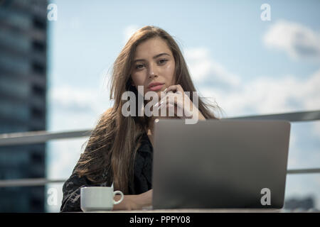 Junge Frau sitzt auf der Terrasse mit Blick auf die Stadt und verfügt über ein Notebook. Stockfoto