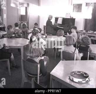 1960, Schule Kinder sitzen mit Percussion Instrumente in einem Musik klasse, mit einer Lehrerin Klavier spielen, England, UK. Stockfoto