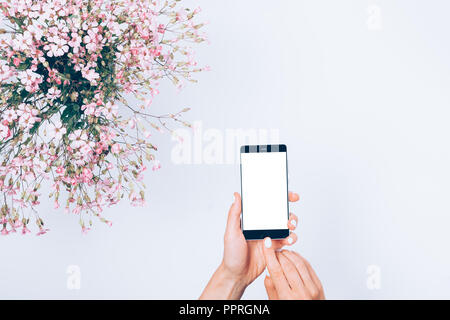 Handy in der Frau die Hände auf weißen Tisch neben dem festlichen Blumenstrauß aus rosa Blüten, Ansicht von oben. Mädchen berühren Bildschirm von smart Gerät mit ihrem Index fi Stockfoto