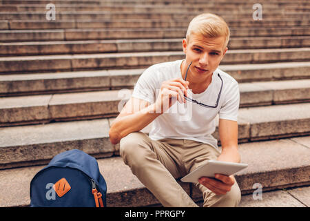 Student mit Rucksack mit Tablet sitzen auf der Treppe und halten Gläser. Guy studieren im Freien. Bildung Konzept Stockfoto