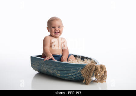 Ein lächelndes sieben Monate alten Baby boy in einem rustikalen, hölzerne Schüssel sitzen. Im Studio auf einem weißen, nahtlose Kulisse erschossen. Stockfoto