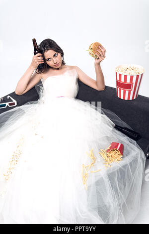 Deprimiert junge Braut im Brautkleid auf der Couch sitzen mit dem Bier und Junk Food auf Weiß Stockfoto