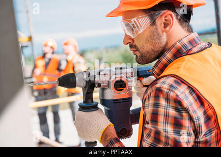 Seitenansicht des Builder in Schutz Helm und Brille mit Bohrmaschine auf der Baustelle Stockfoto