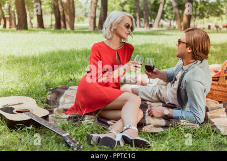 Glückliches Paar liegen auf Decke in Park und klirren mit Gläser Rotwein Stockfoto