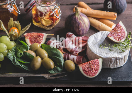 Prosciutto, Camembert, Feigen, Spinatblätter, Honig, Trauben, Oliven auf dunklen, das Brett über rustikal Hintergrund Stockfoto