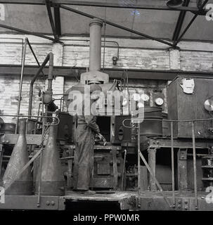 1950 s, historischen, einen männlichen Arbeitnehmer in Latzhosen und Baskenmütze stehend an einer industriellen Ofen bei einer Reifenfabrik. Stockfoto