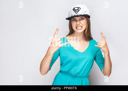 Portrait von kühlen aggressiver junge schöne Mädchen im blauen Kleid und weiße Kappe mit Übermenschen Symbol stehend erhobenen Armen, anzeigen Rock unterzeichnen und Zähne. In Stockfoto