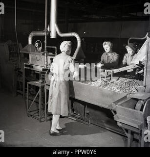 1950, historische, weibliche Fabrikarbeiter in Overalls und Haar Schals stehen Fächer der metallischen Komponenten von Maschinen Hersteller Zink-Kohle-Batterien, stets bereit, Wald Rd, Walthamstow, London, England, UK produziert. Stockfoto