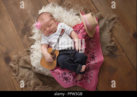 Ein 9 Tag alt, sleeing Baby Mädchen in Jeans und halten eine kleine akustische Gitarre. Sie liegt in einer Holzkiste mit Lammfell und einem rosa Verbot gefüttert Stockfoto