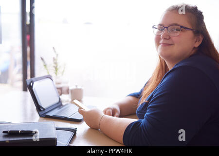 Chubby Geschäftsfrau tragen ein Navy Bluse und Gläser, an der Kamera, während in einem Café mit einer Vielzahl von Technologie vor ihr sitzt auf der t Stockfoto