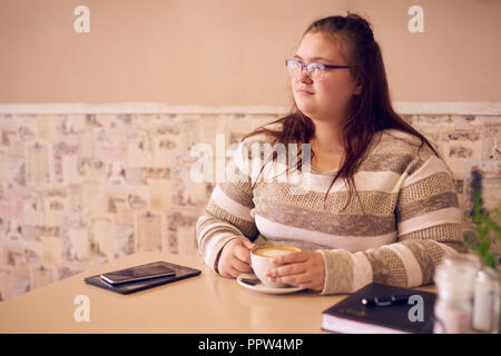 Übergrößen Frau sitzt in einem hellen Café, während Sie bei einer Tasse Kaffee ihre Hände warm halten und bereit mit Ihrem Tablet-PC auf dem Tisch weiter arbeiten können. Stockfoto