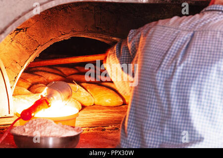 Der Bäcker nimmt ein Laib Brot aus einem alten Backofen mit einem hölzernen Löffel. Reihen von frisch gebackenen Brötchen Stockfoto