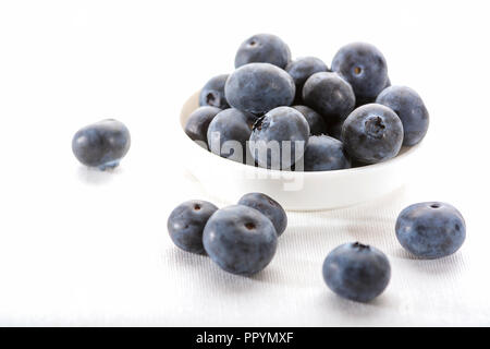 Nahaufnahme von einer weißen Schüssel mit saftigen Blaubeeren auf einer weißen Tischdecke mit einigen blueberrie auf dem Tisch verstreut Stockfoto