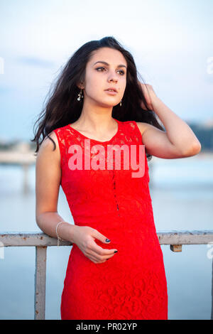 Schöne junge Mädchen im roten Kleid auf einer Brücke in der Nähe einer alten Zaun posieren. Im Hintergrund gibt es einen Fluss, Brücken und Fragmente der städtischen Gebäude Stockfoto