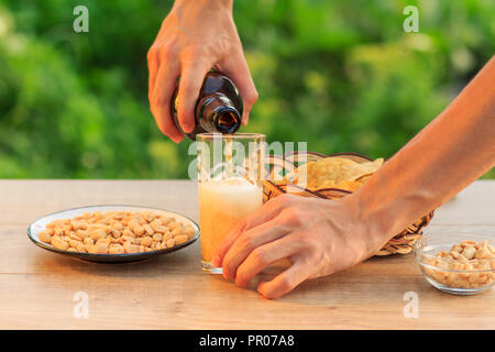 Junger Mann hält eine Flasche Bier und füllen das Glas. Männliche hand Bier ins Glas auf Holztisch mit Kartoffelchips in Weidenkorb gießen, peanu Stockfoto