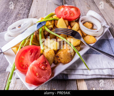 Kartoffeln gebraten mit weißen Zwiebeln und Tomaten auf, in eine weiße Platte, auf alten Holztisch, rustikaler Stil, Nahaufnahme. Stockfoto