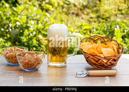 Glas Becher Bier auf Holztisch mit Kartoffelchips in Weidenkorb, Erdnüsse und getrockneten Tintenfisch in Schalen auf natürlichem Grün verschwommenen Hintergrund Stockfoto