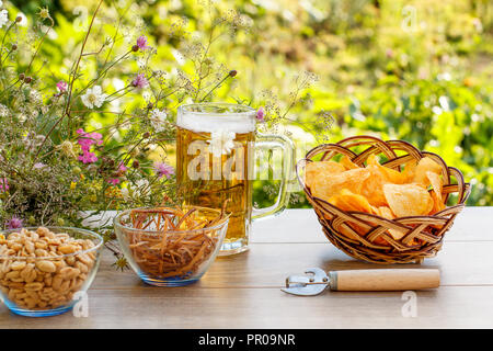 Glas Becher Bier auf Holztisch mit Kartoffelchips in Weidenkorb, Erdnüsse und getrockneten Tintenfisch in Schalen auf natürlichem Grün verschwommenen Hintergrund Stockfoto