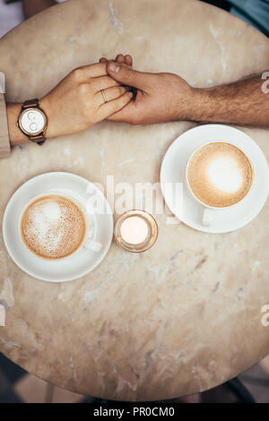 Ansicht von oben in die Hände von Mann und Frau zusammen im Ausdruck der Liebe. Der Mann die Hand einer Frau, die auf einem Tisch mit Tassen Kaffee durch die Stockfoto