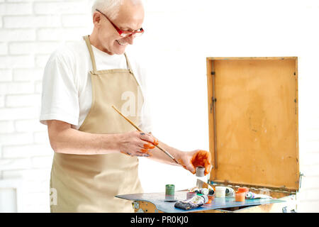 In der Nähe von älteren gutaussehenden Mann Auswahl malt er braucht neue Malerei in helles Studio zu malen. Professional Master tragen in Schürze und in Gläsern, in schmutzige Farbe der Hände, die Pinsel, lächelnd. Stockfoto
