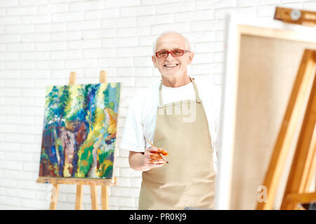 Kreative senior Maler Zeichnung farbenfrohe abstrakte Malerei an helles Studio. Schöner Mann, tragen, Schürze und Gläser, lächelnd und mit Blick auf die Kamera mit fertigen Bild auf verschwommenen Hintergrund. Stockfoto