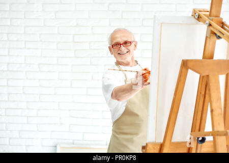 Gut aussehender älterer Maler Zeichnung farbenfrohe Malerei an helles Studio. Glücklich, Mann, tragen, Schürze und Gläser, lächelnd und Suchen von staffelei mit Bild in der Kamera mit und Veredelung Meisterwerk. Stockfoto