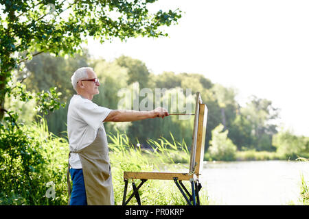 Seitenansicht des stattlichen männlichen Maler Zeichnung Bild am Tag im Park mit See. Senior Artist, trug in Schürze und moderne Gläser, mit der Arbeit an seinem neuen Bild, mit Palette mit Farben und Pinsel. Stockfoto