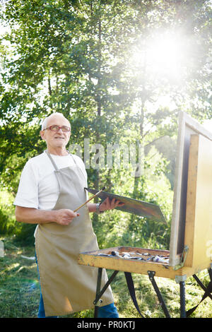 Stattliche männliche Künstler, mit Begriffen, die Bürste und Vorstellen Bild im Kopf in den Strahlen der Sonne. Senior Maler, tragen, Schürze und moderne Gläser, mit Palette mit Farben gegen See. Stockfoto