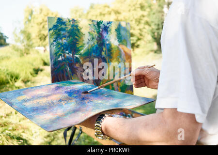 Close-up Künstler Hand Malerei Pinsel und Palette der Farben gegen fast fertige Bild im Park. Staffelei und Leinwand mit Bild stehend gegen See auf Open Air. Konzept der Hobby. Stockfoto