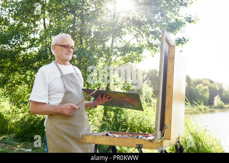 Ältere männliche Maler, stehend mit Malerei Pinsel und Palette von Farben in der Hand, Kommissionierung bis Farbe Bild im Strahlen der Sonne. Gutaussehenden Mann im weißen T-Shirt und Schürze, bei der Arbeit im Park. Stockfoto