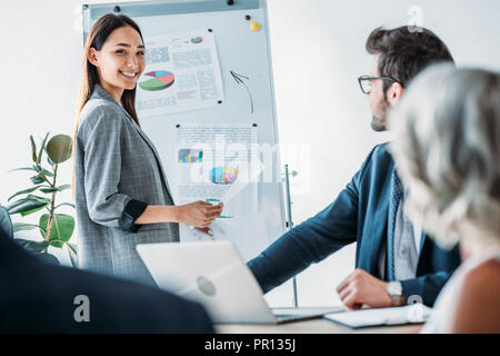 Lächelnd asiatischen Geschäftsfrau, die in der Nähe von Flipchart während der Sitzung im Büro und suchen an Kamera Stockfoto