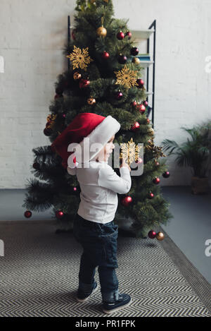 Nett lächelnden Jungen in Santa hat an Weihnachten Baum zeigen Stockfoto