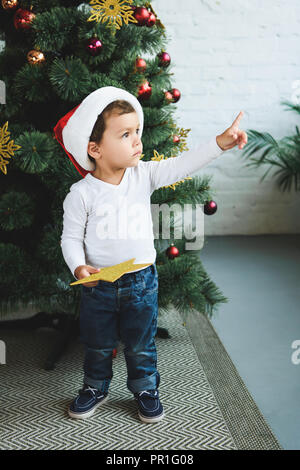 Adorable Junge in Santa Hut und goldene Sterne, während nach oben zeigt, und in der Nähe von Weihnachten Baum Stockfoto