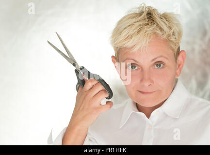 Reife Frau mit kurzen, blonden Haare, in einem weißen Hemd ist hält geöffnete Schere in ihren Händen, sie lächelt, einen hellen Hintergrund Stockfoto