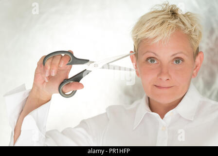 Reife Frau mit kurzen, blonden Haare, in einem weißen Hemd ist hält geöffnete Schere in ihren Händen, sie lächelt, einen hellen Hintergrund Stockfoto