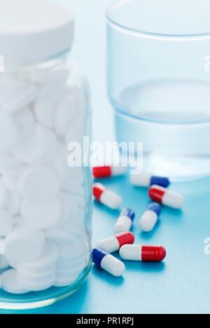 Weiße Pillen in ein Glas Flasche und ein Glas Wasser mit verschiedenen anderen Droge Kapseln im Hintergrund. Stockfoto