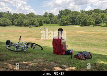 Nicht identifizierbare Person ein Buch lesen in Piedmont Park an einem sonnigen Tag mit Fahrrad und Rucksack und andere Menschen spielen in der Ferne auf dem Grün gr Stockfoto