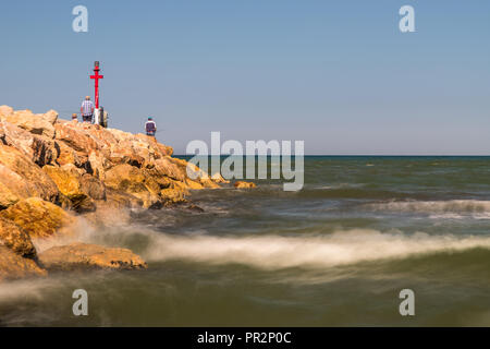 Goldene Stunde lange Exposition der Wellen auf die Felsen, wo einige Fischer in der Nähe von einem roten Bootfahren Markierung stehen. Stockfoto