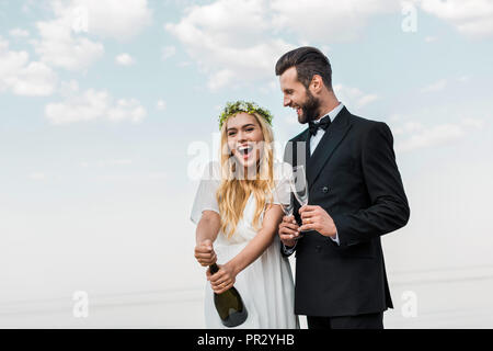 Glückliche Braut im weißen Kleid öffnen Flasche Champagner am Strand Stockfoto