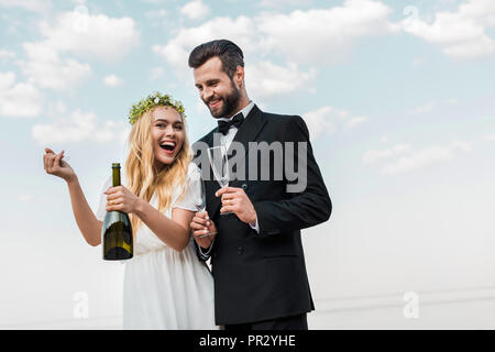 Lachen Braut im weißen Kleid öffnen Flasche Champagner am Strand Stockfoto