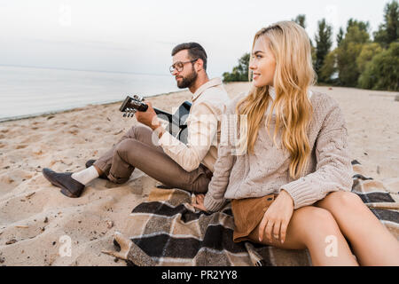 Schöner Freund im Herbst Outfit spielen akustische Gitarre für Freundin am Strand. Stockfoto