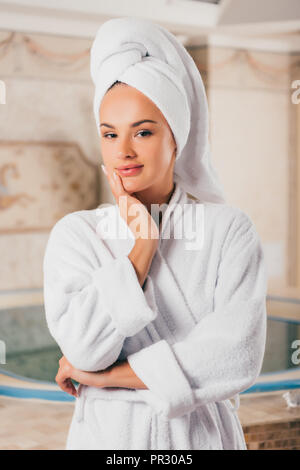 Lächelnde Frau in weißem Frottee Bademantel und Handtuch auf dem Kopf im Spa Center mit Schwimmbad Stockfoto