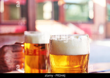 Detaillierte Foto von zwei Gläser Bier mit weißer Schaum in einem Gartenrestaurant mit einem unscharfen Hintergrund und Sonnenlicht Stockfoto