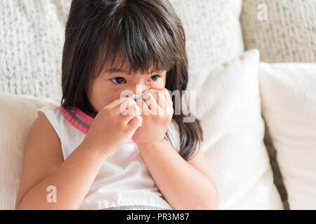 Kranken kleinen asiatischen Mädchen wischen oder Reinigung der Nase mit einem Taschentuch, auf dem Sofa zu Hause sitzen. Medizin und Gesundheit Konzept. Stockfoto